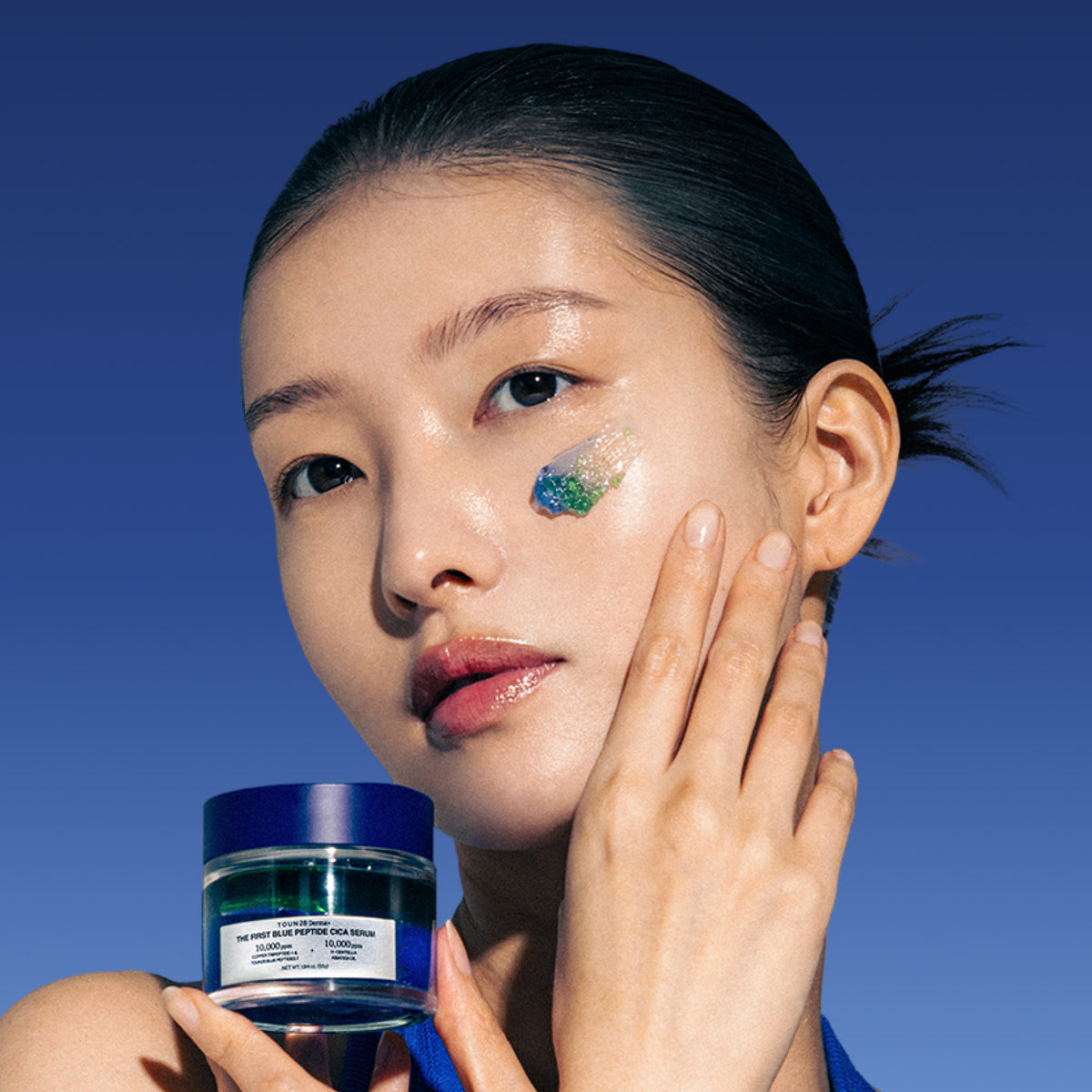 Model holding 1% Blue Peptide Cica Serum with blue and green serum texture on her cheek