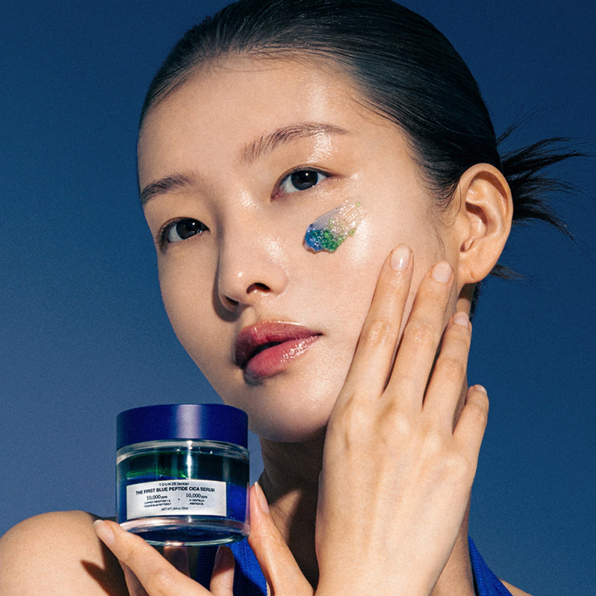 Model holding 1% Blue Peptide Cica Serum with blue and green serum texture on her cheek